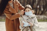 Crop female helping to put on medical mask for daughter during stroll in nature