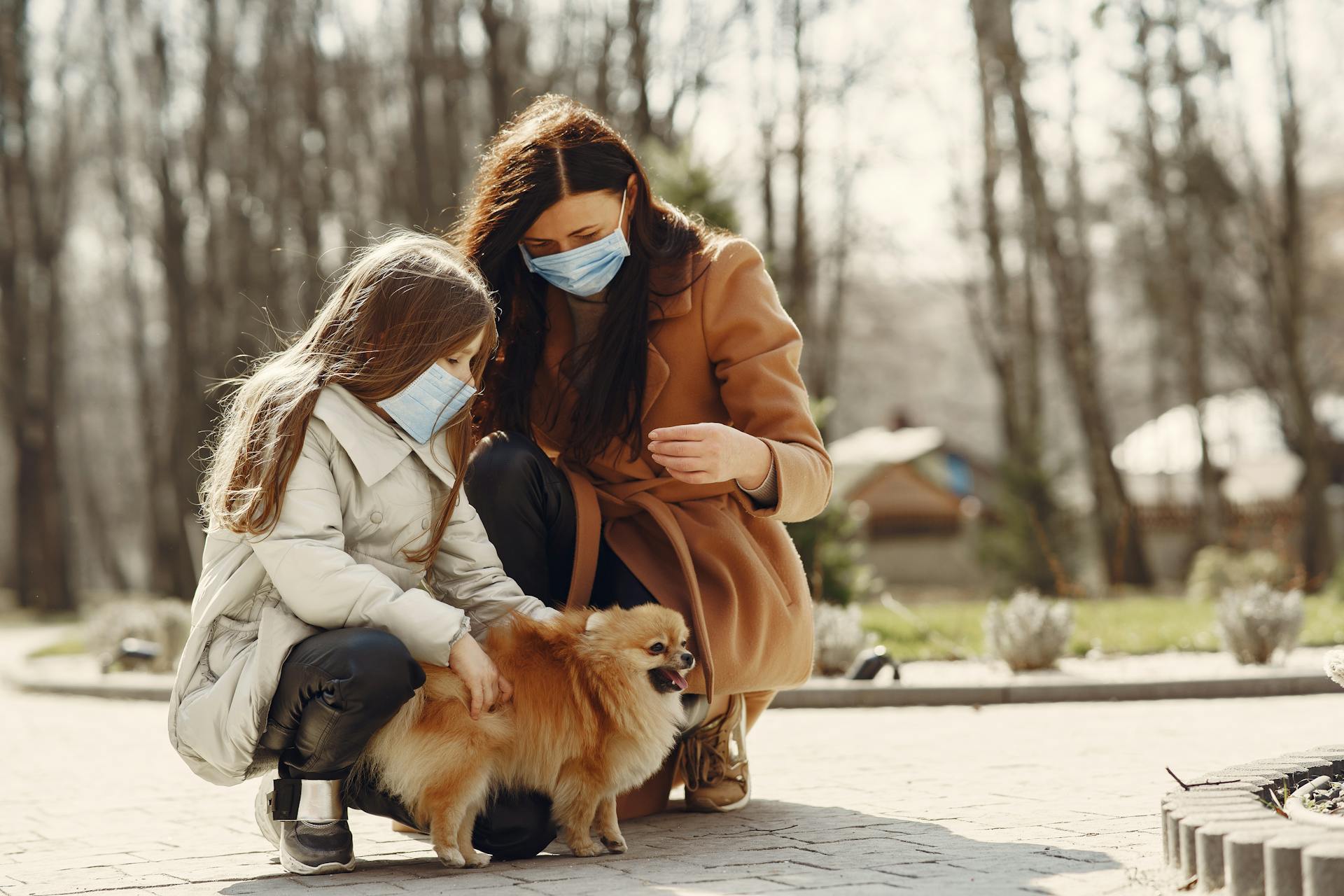 Cheerful daughter and mom in warm clothes and medical masks with little Pomeranian dog enjoying weekend in nature together