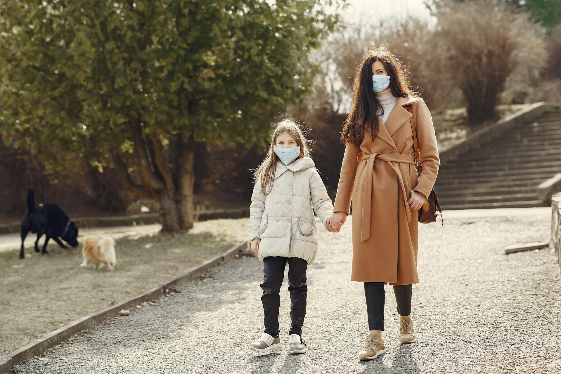 Woman with long hair in face mask holding hand of small girl while walking in park