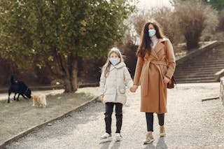 Woman with long hair in face mask holding hand of small girl while walking in park