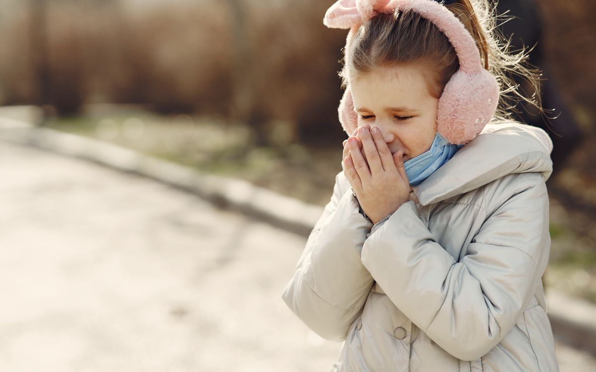 Small child with lowered face mask covering face by hands while sneezing on street
