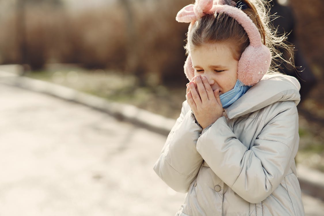Small child with lowered face mask covering face by hands while sneezing on street