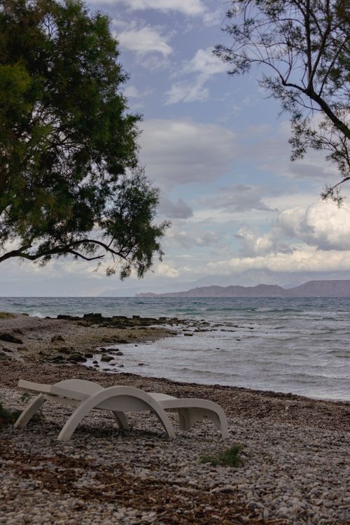 Free Serene rocky beach with lounge chairs under trees and a calm sea view, perfect for relaxation. Stock Photo