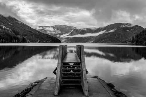 Free A black and white image of a tranquil lake with a wooden jetty and misty mountains in Norway. Stock Photo