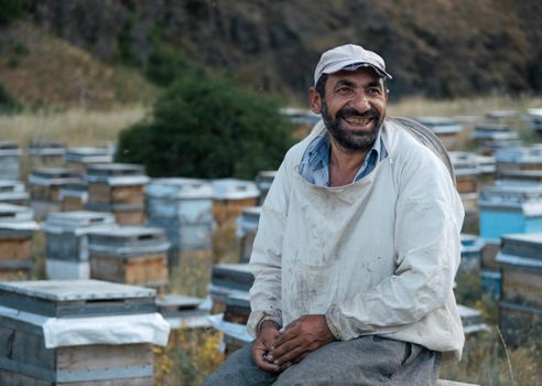 A cheerful beekeeper in protective clothing sits among wooden hives in a rural setting.