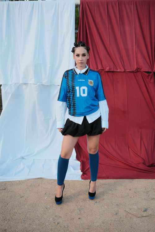 Free Model poses in a stylish soccer jersey against a red and white backdrop outdoors. Stock Photo