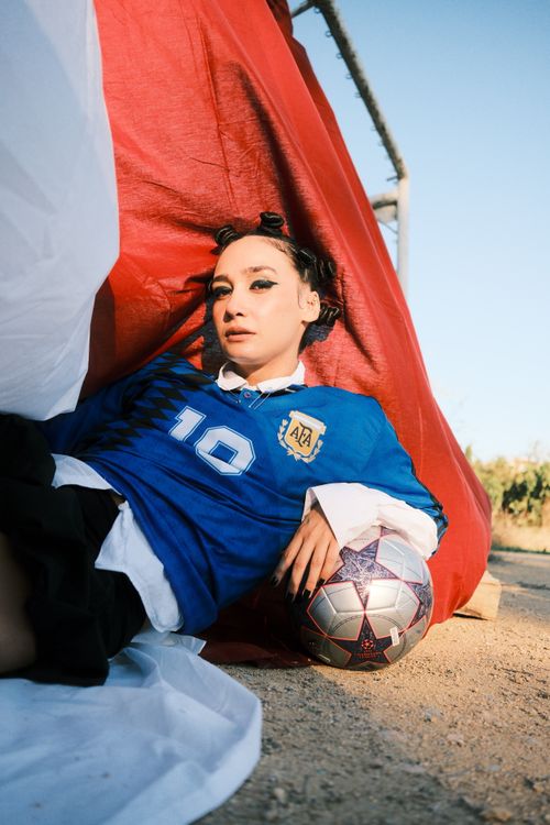 Free Woman in blue soccer jersey posing with a ball against a red backdrop outdoors. Stock Photo