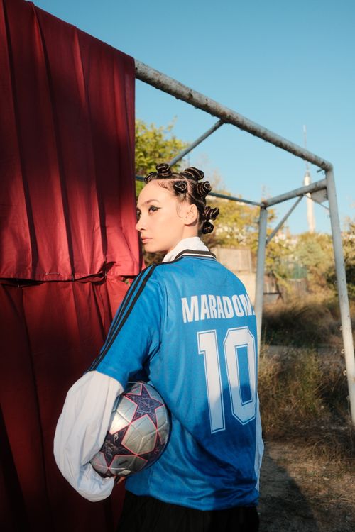 Free Young person with stylish hair in a Maradona jersey by a goal post, holding a soccer ball outdoors. Stock Photo