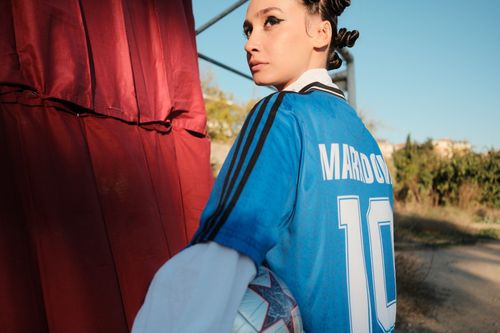 Free A woman in a Maradona jersey gazes back while holding a soccer ball in an outdoor setting. Stock Photo