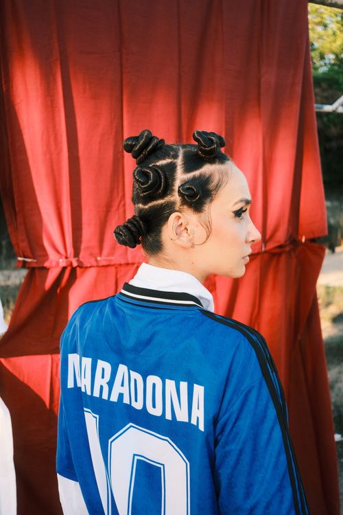 Free A woman in a blue Maradona jersey with unique hair poses outdoors against a vibrant red backdrop. Stock Photo
