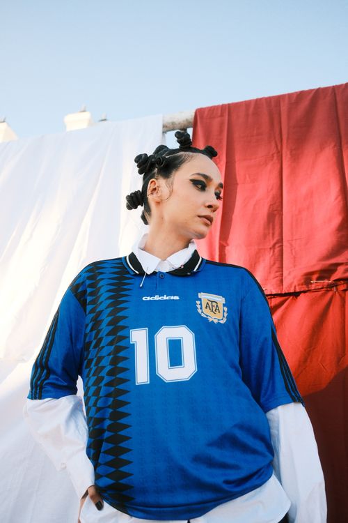 Free Woman wearing a blue soccer jersey poses confidently outdoors against a red and white backdrop. Stock Photo