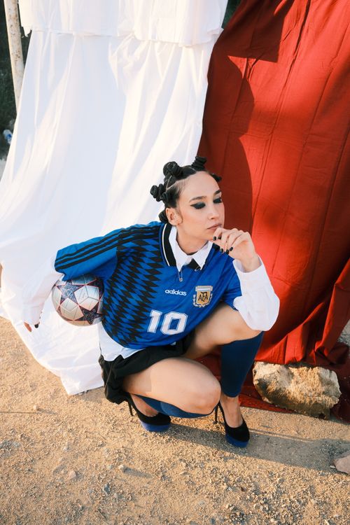 Free A stylish woman in a soccer jersey crouches outdoors with a football. Stock Photo