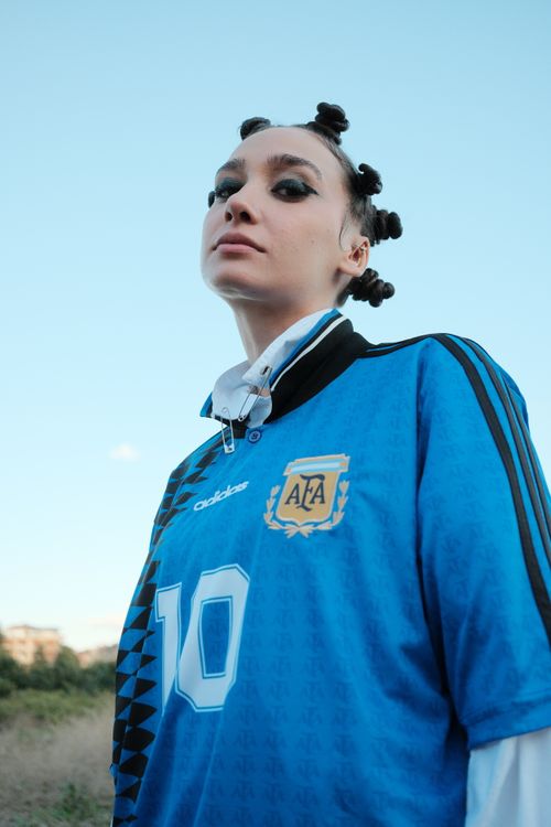 Free Woman poses confidently in a blue soccer jersey with a striking hairstyle against a sky backdrop. Stock Photo