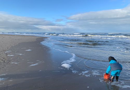 Free A child in winter clothing plays on a sandy beach with ocean waves in the background. Stock Photo