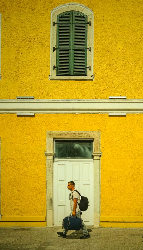 Free Man with luggage walking by a bright yellow building, showcasing travel and urban life. Stock Photo