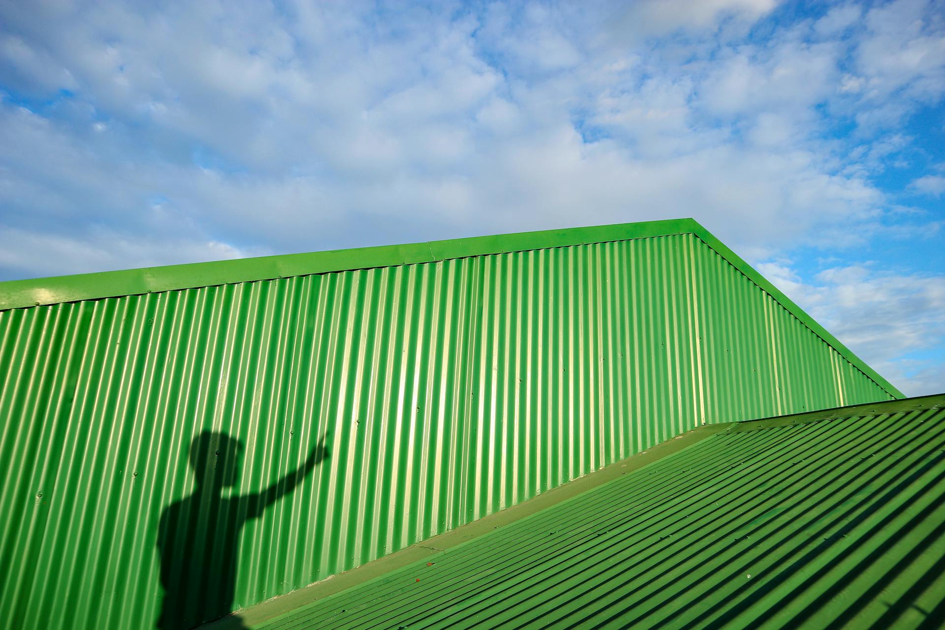 Green corrugated metal structure with a person's shadow against a blue sky.