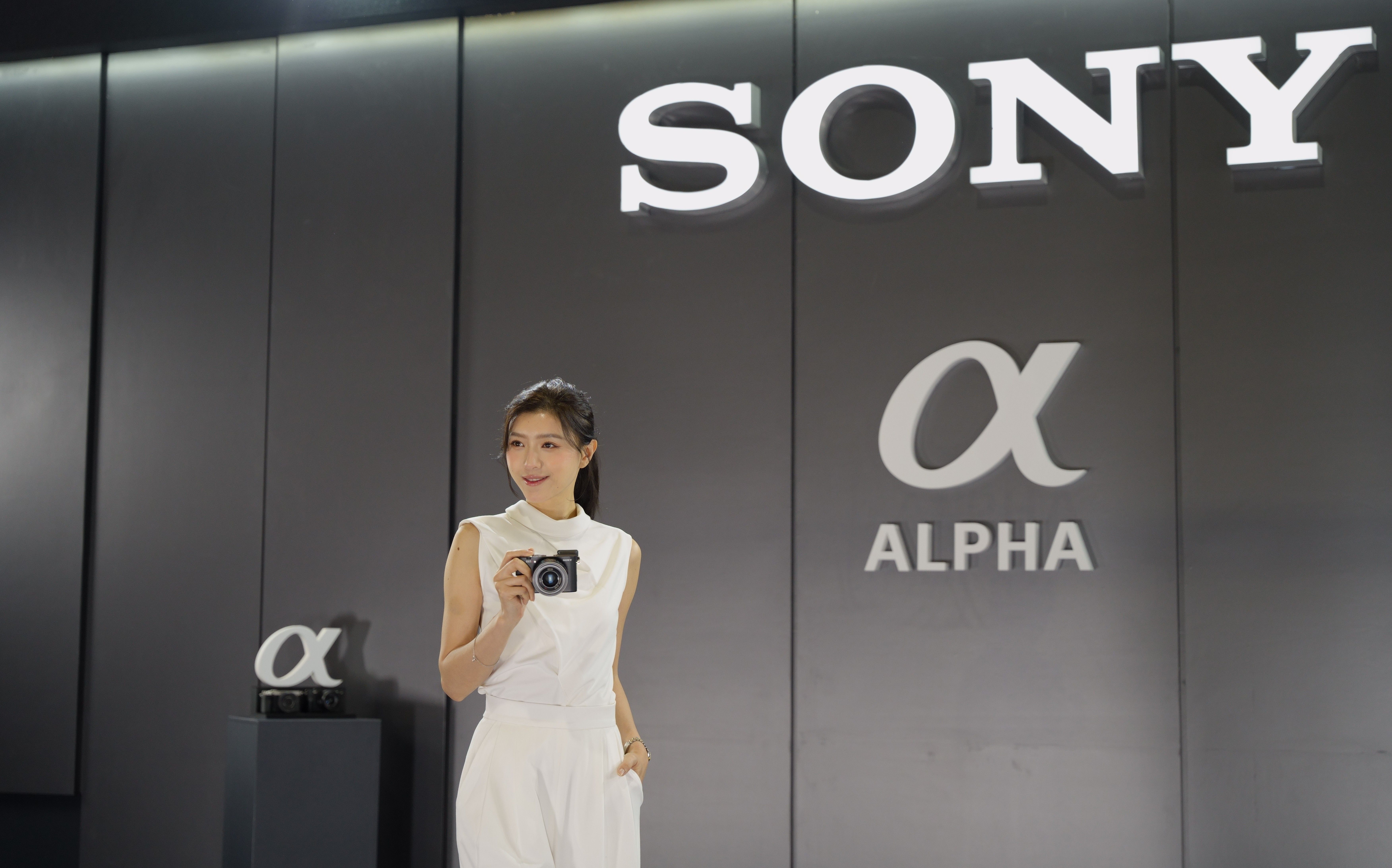Woman in white, holding a camera, stands at a photography event under large logo signage.