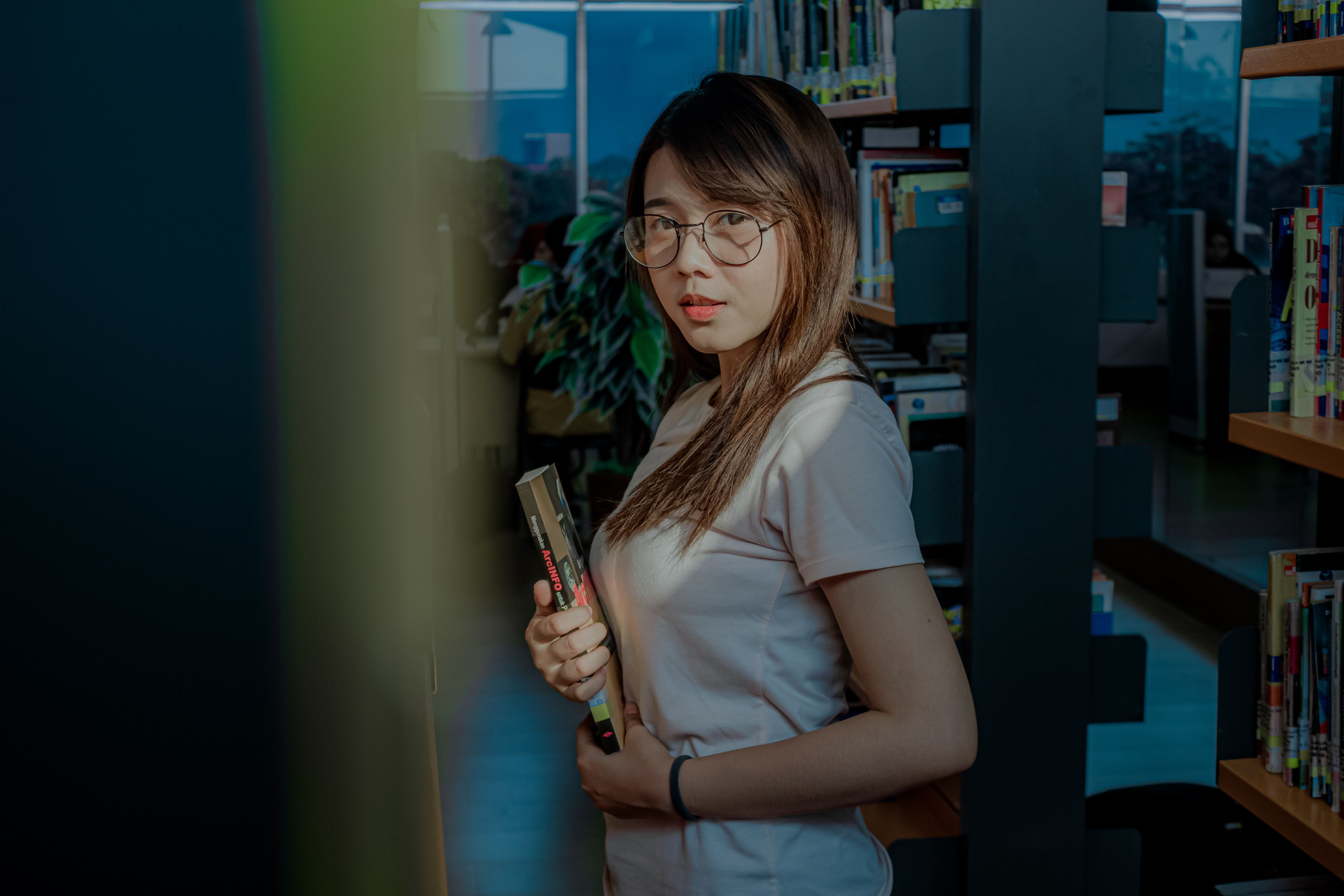 Woman standing in library aisle holding book, with soft natural lighting creating a calm atmosphere.