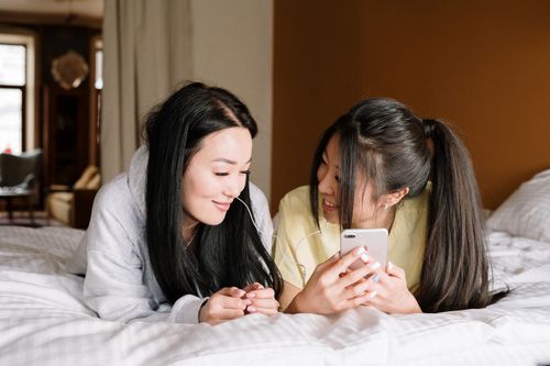 Free Two friends enjoying a cozy day at home, sharing earphones and using a smartphone on the bed. Stock Photo