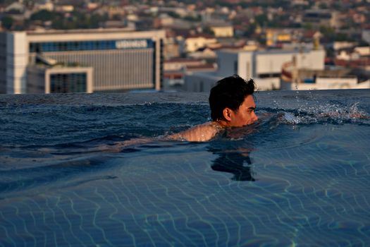 An adult male swimming in an infinity pool with a panoramic urban skyline in the background.