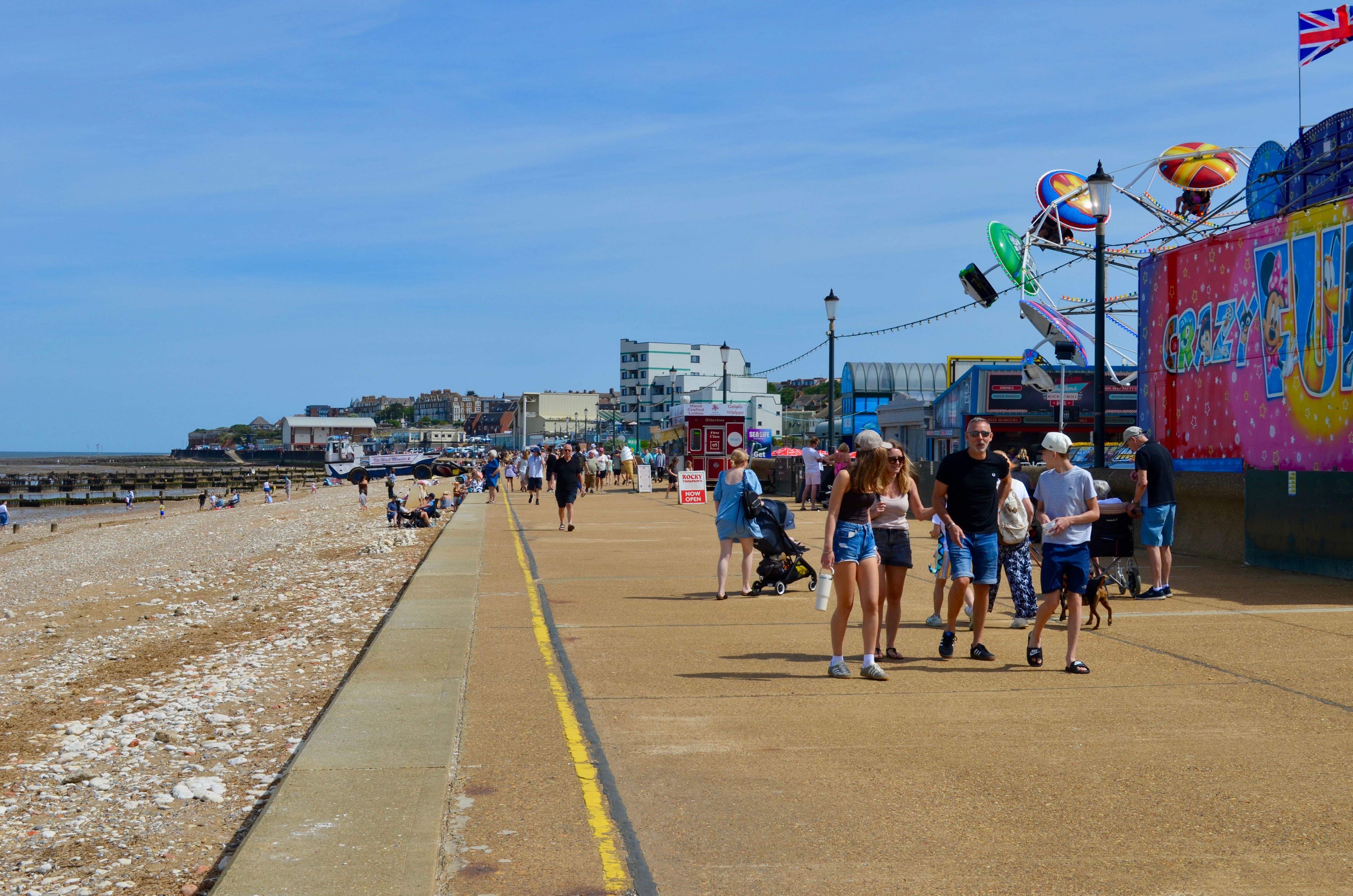 People enjoying a sunny day at the seaside promenade with a lively amusement area and clear blue skies.