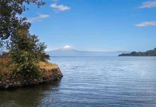 Peaceful lake with lush greenery and distant snow-capped volcano under a clear blue sky.
