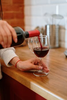 A person pouring red wine into a glass in a warm, cozy kitchen setting.