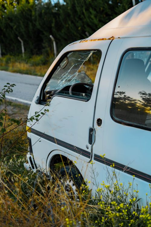 Free A broken-down white van with a shattered window sits abandoned by a rural roadside, captured in late afternoon light. Stock Photo