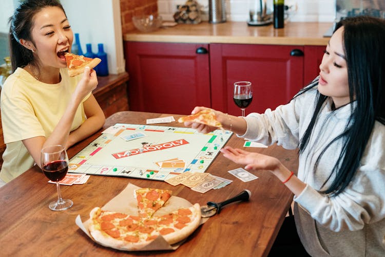 Women Snacking On Pizza While Playing Monopoly 