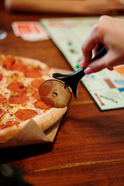 Free Hand holding a pizza cutter slicing into pepperoni pizza next to a board game on a wooden table. Stock Photo