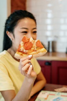 A young woman smiles while holding a piece of pepperoni pizza in her cozy home kitchen.