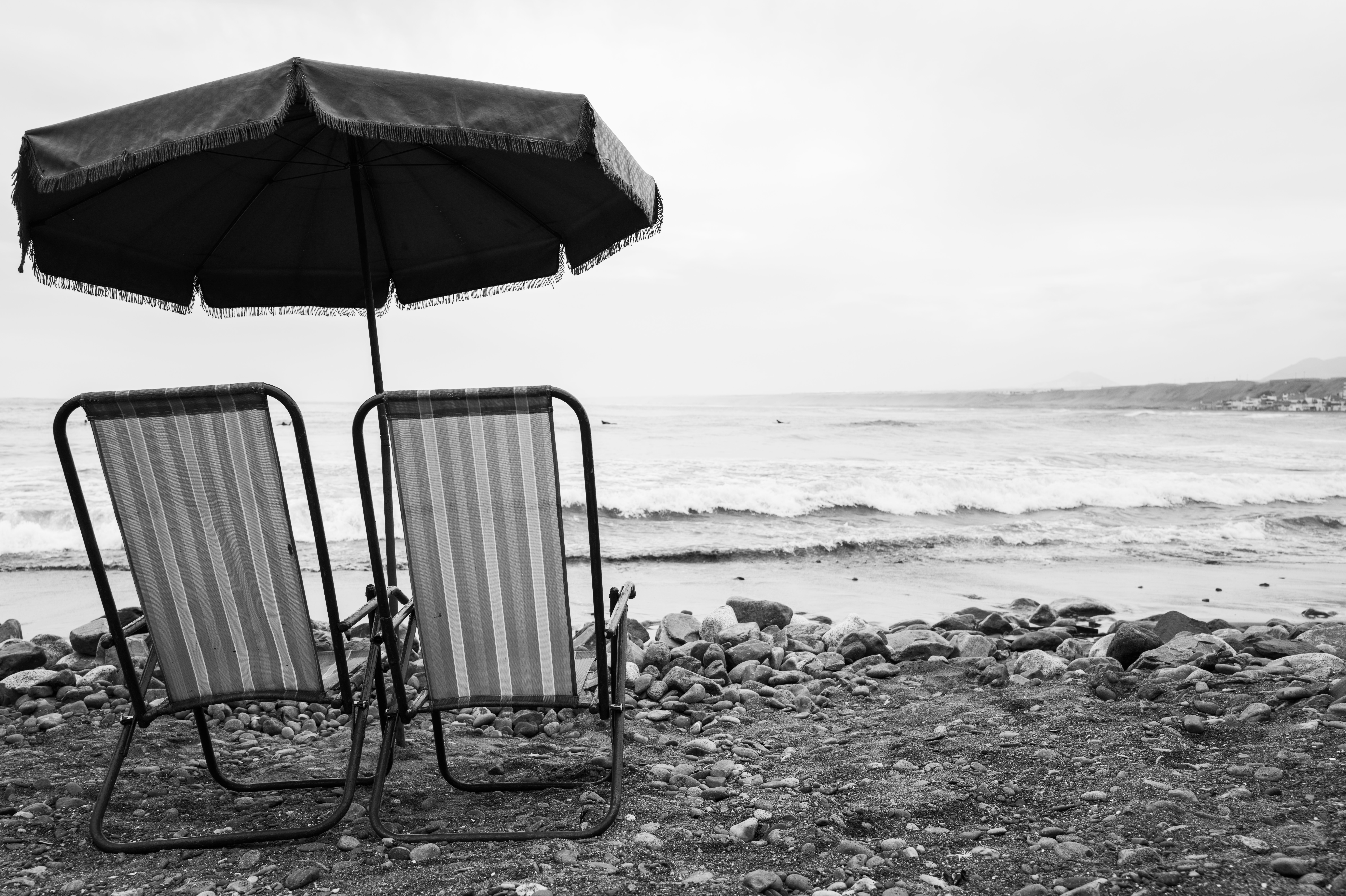 Two beach chairs under an umbrella on a rocky shore in black and white.