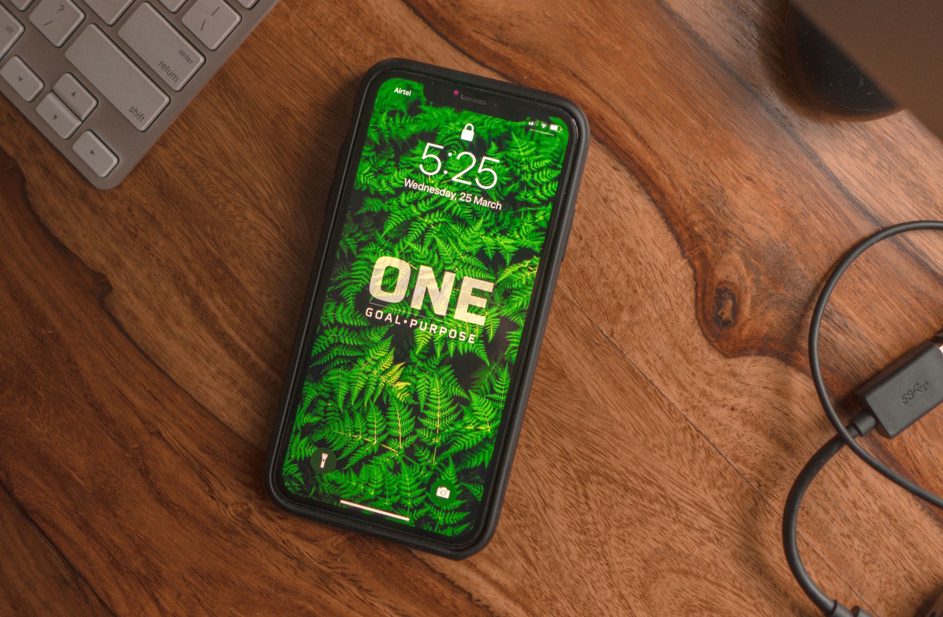 A smartphone with a green fern wallpaper and motivational text on a wooden desk, alongside a keyboard.