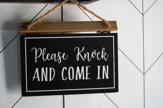 A black, wooden door sign reading please knock and come in.