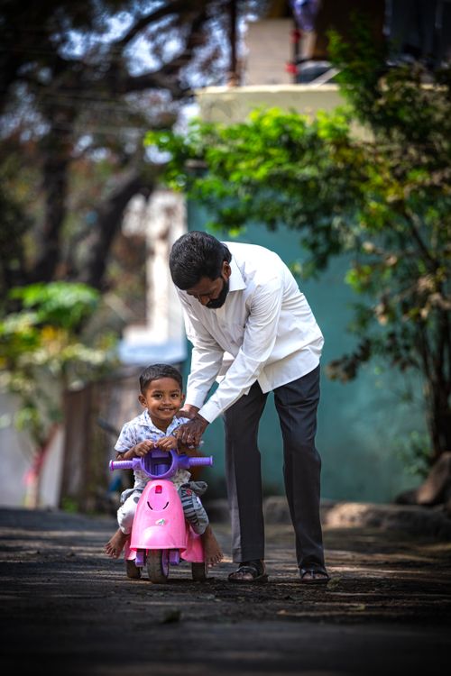 Free A father assists his young son in riding a pink toy motorcycle outside on a sunny day. Stock Photo