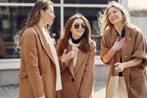 Cheerful female friends with long hair in elegant coats smiling while happily chatting with each other on street