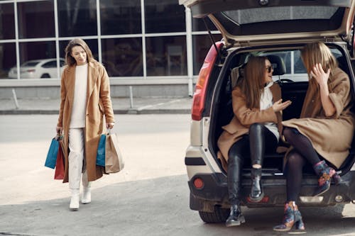 Free Cheerful females with long hair in elegant coats cheerfully talking with each other while waiting for friend coming after shopping Stock Photo