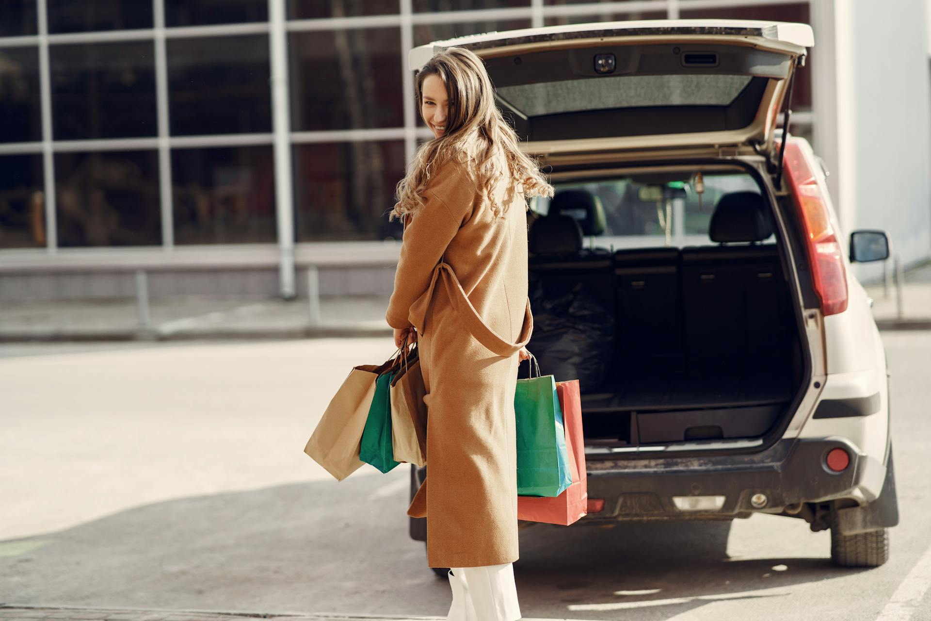 Full length of cheerful female looking over shoulder while standing near opened trunk of car on parking of shopping mall