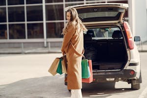 Full length of cheerful female looking over shoulder while standing near opened trunk of car on parking of shopping mall
