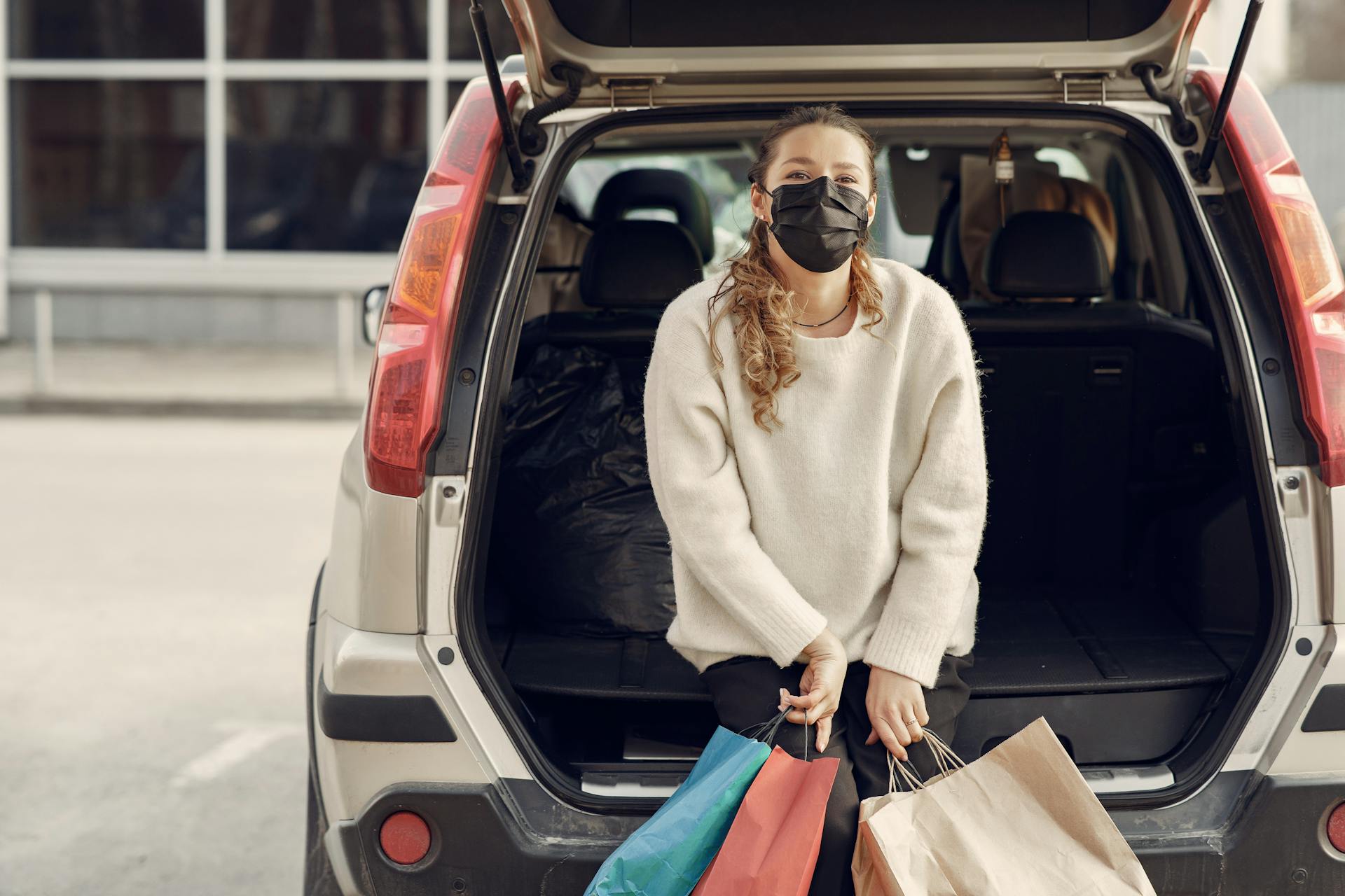 Young woman in casual clothes and protective mask sitting in trunk of modern car holding shopping bags with purchases on city street