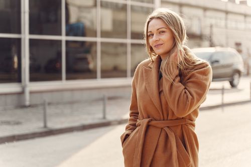 Free Positive female in stylish warm coat standing on city street against modern glass building and touching hair in back lit Stock Photo