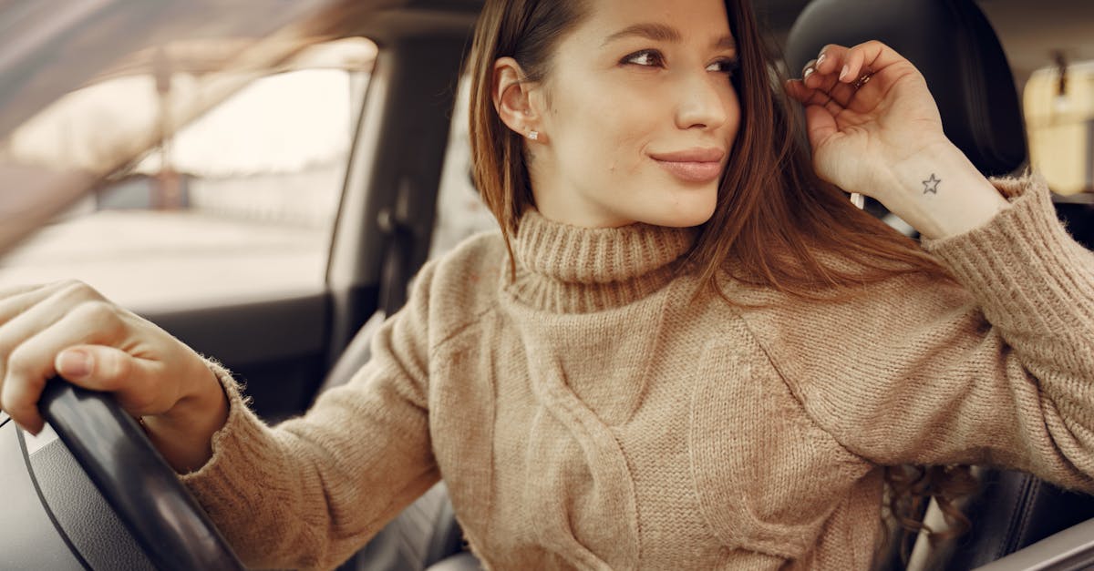 Positive young female driver in warm clothes at wheel of leather interior car going around city streets while greeting friend during riding