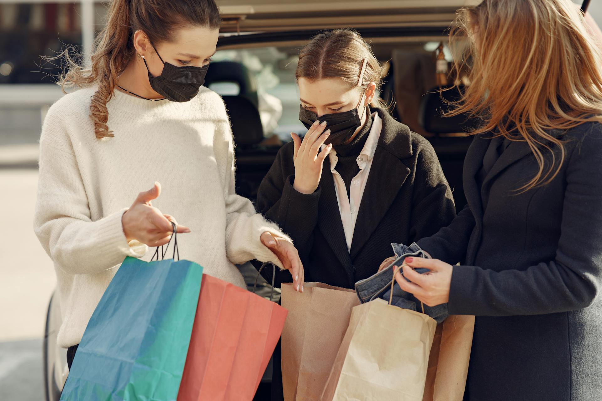 Crop women demonstrating purchases in shopping bags to each other standing in protective medical masks near opened trunk of car on street