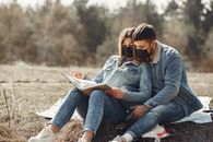 Young couple in jeans clothes and protective masks sitting on blanket in field and looking at photo album in back lit