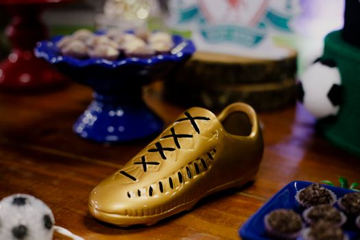 Bright artificial sneaker with shoelaces on wooden desk near plate and stand with delicious cakes and treats near decorative soccer ball in flat during celebration