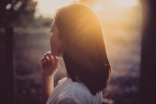 Side view of unrecognizable wistful ethnic female touching chin while standing under bright sunshine in rural zone in back lit in evening