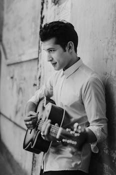 A stylish young man playing guitar against a rustic wall in monochrome.