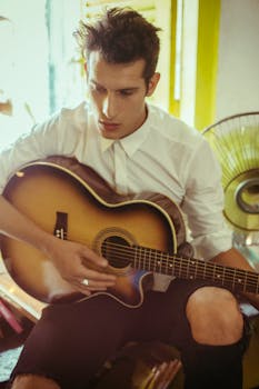 A young man plays an acoustic guitar inside a cozy room, wearing casual attire.