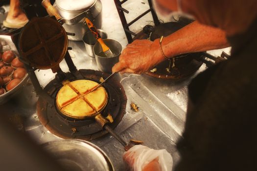 An elderly person prepares waffles using a vintage waffle iron at a street food stall.