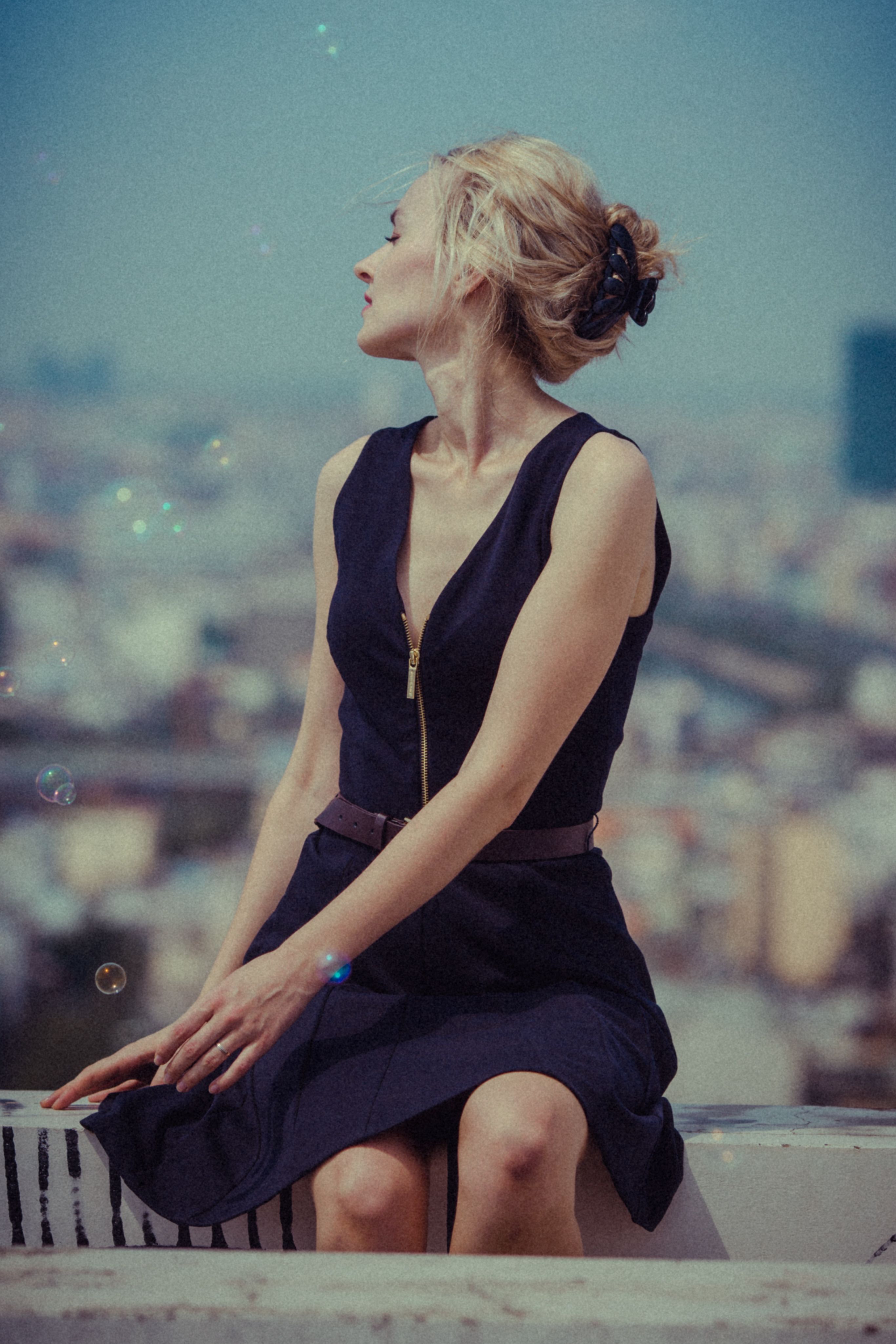 Blonde woman sitting in black dress, overlooking a city skyline on a sunny day.
